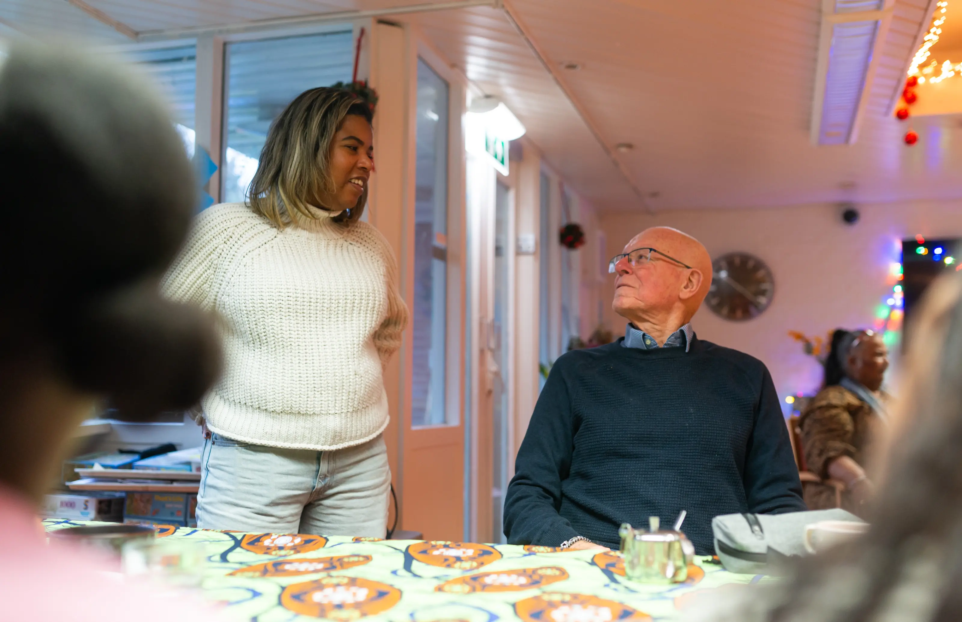 Een oudere man zit aan een tafel en kijkt naar een jonge vrouw die met hem in gesprek is. Dit laat zien hoe jongeren en ouderen binnen het project met elkaar in contact komen en samen een fijne tijd hebben.