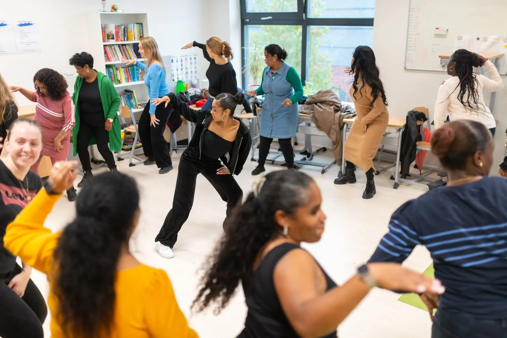 Een diverse groep vrouwen neemt deel aan een energieke dansworkshop in een klaslokaal, geleid door een enthousiaste instructeur. De workshop maakt deel uit van een educatief programma van Stichting Eduvibes, gericht op talentontwikkeling en persoonlijke groei voor jongeren en volwassenen.